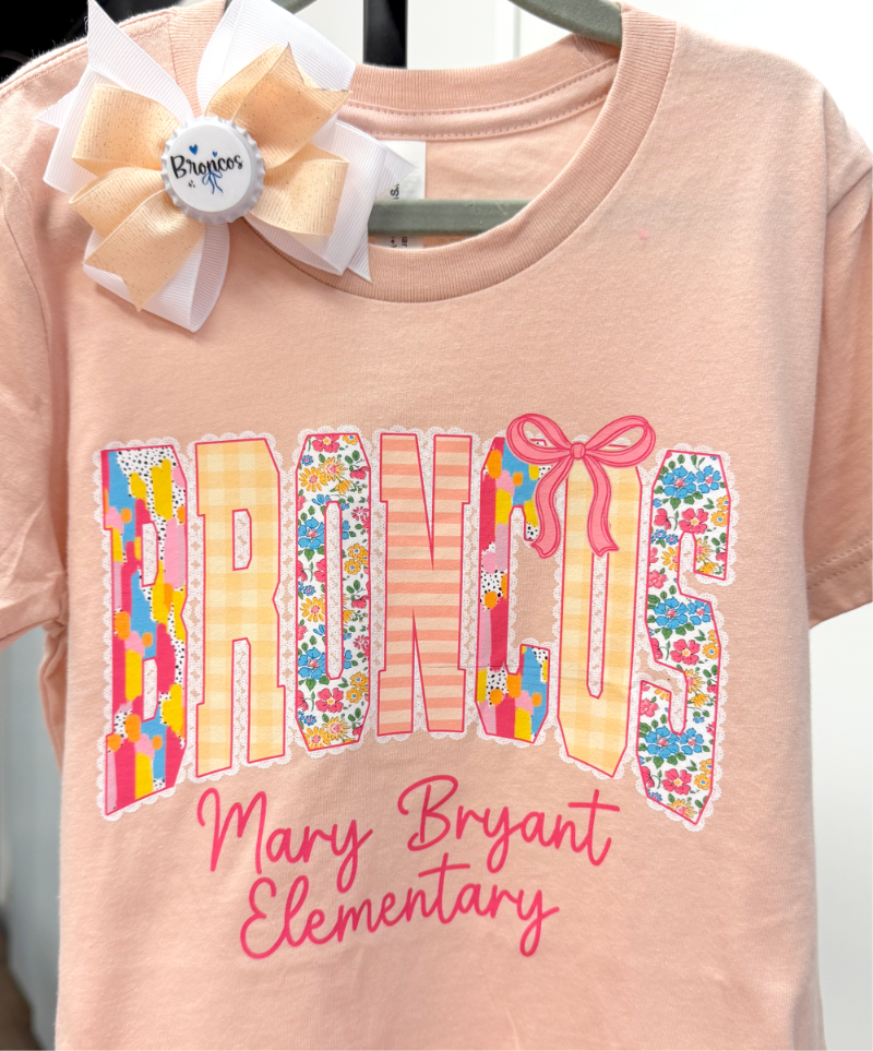 A light peach-pink short-sleeve t-shirt featuring a large 'BRONCOS' graphic with various patterned fillings and a pink bow detail, with 'Mary Bryant Elementary' written below in pink script. A large cream and peach hair bow is attached to the collar.